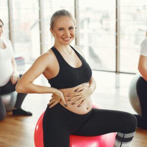 Adult pregnant females in activewear sitting on fit balls while doing exercises with instructor in modern studio of fitness center